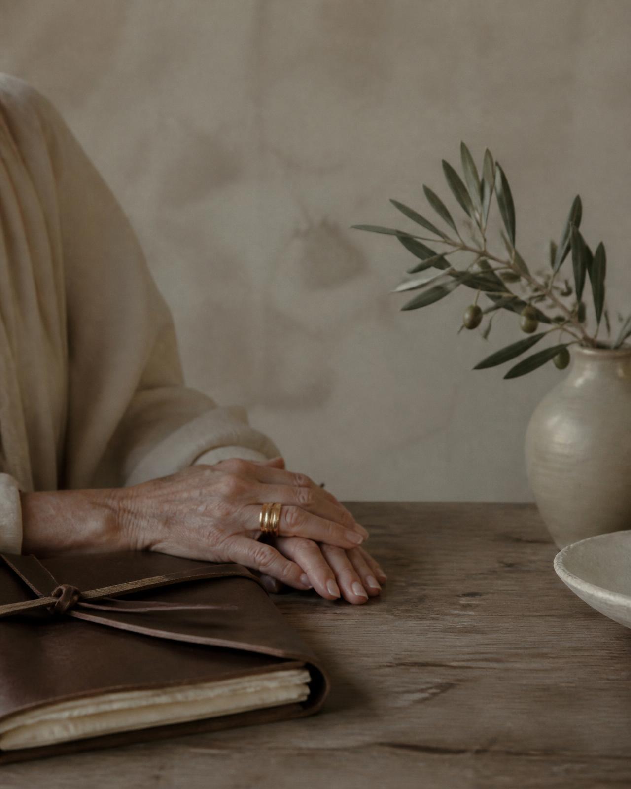 An older woman's hands resting beside a leather journal on a weathered wooden table